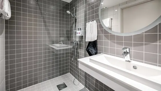 Et moderne badeværelse med flisebelagt bruseområde og spejl over en hvid vask. Blød belysning og neutrale toner skaber et rent, minimalistisk udseende.
