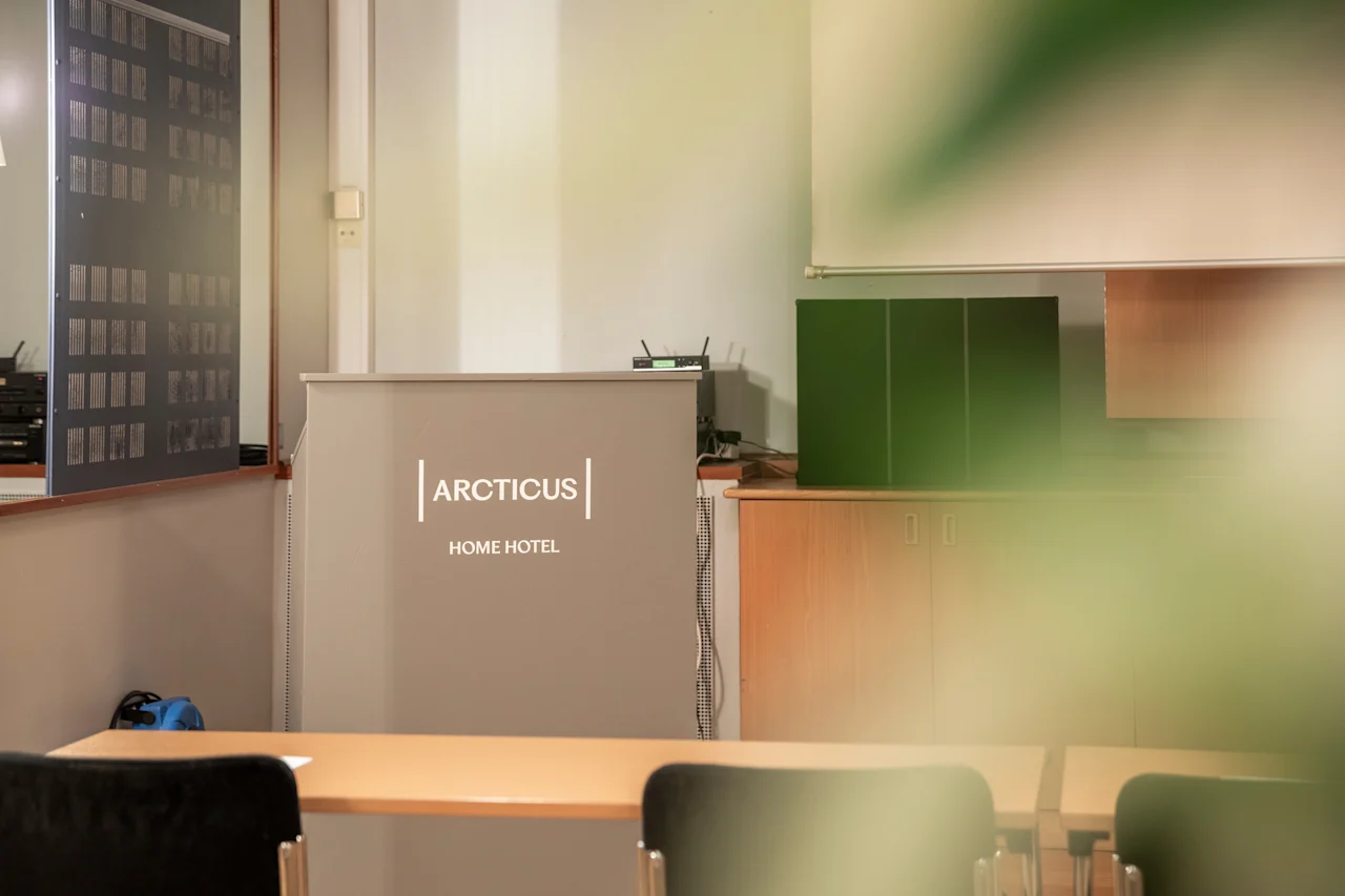 Vastaanottotiski hotellin aulassa, jossa lukee HOME HOTEL. Tuolit ja pöytä ovat etualalla, mikä viittaa odotusalueeseen.