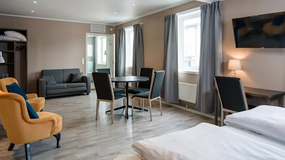 A modern hotel room with a bed in the foreground, a round dining table with chairs, a small sofa, and a wall-mounted TV under recessed lighting.
