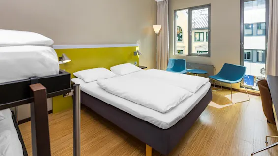 A double bed with white linens stands in a modern hotel room, accompanied by a bunk bed, two blue chairs, and a small table near a window with city views.