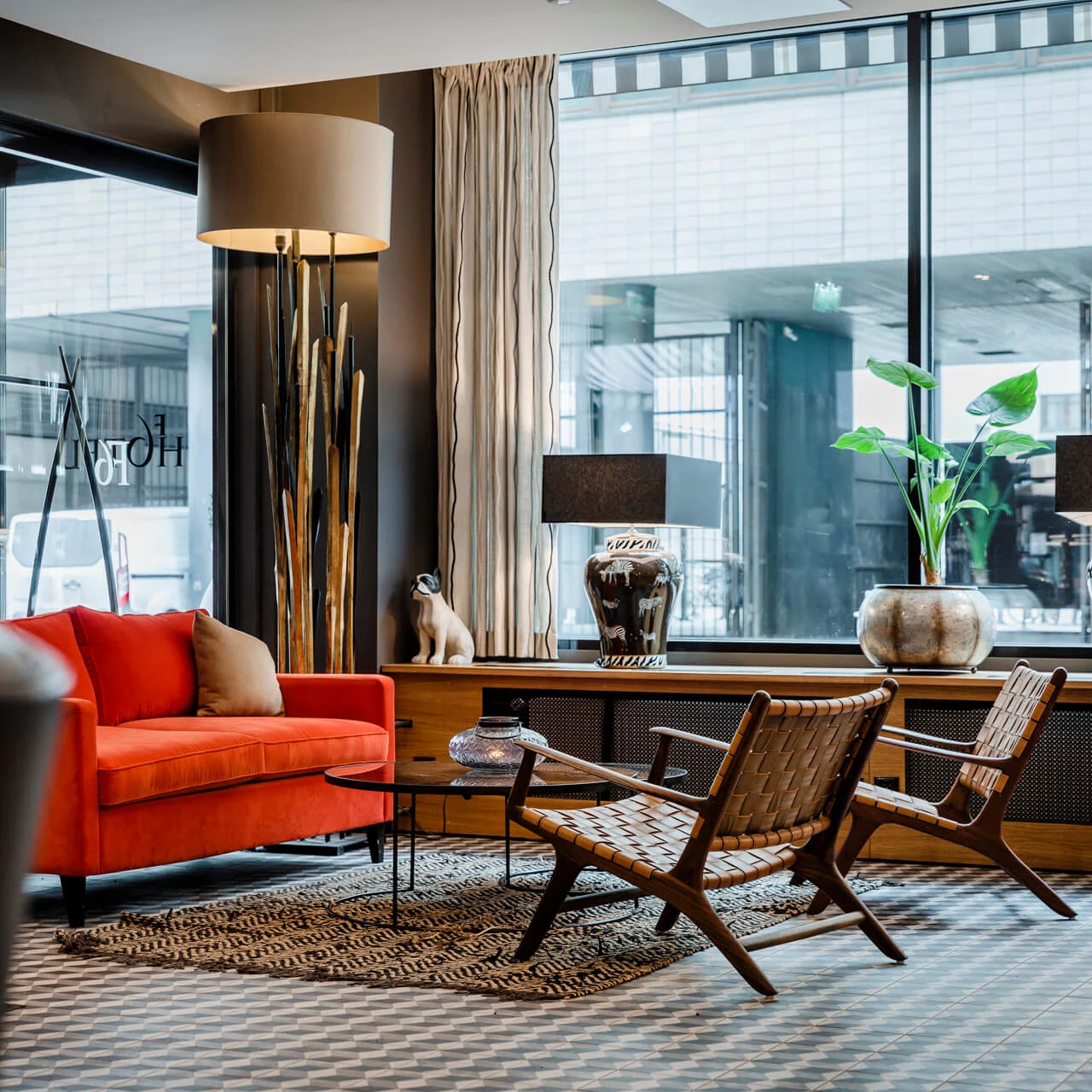 Rød sofa står i et moderne lobbyområde, omgitt av kurvstoler og et rundt bord. Store vinduer viser teksten "HOTEL", med en urban utsikt utenfor.