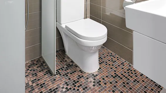En vit toalett är placerad mot beige kakelväggar, bredvid en glasdusch och en vit handfat i ett kompakt, modernt badrum.