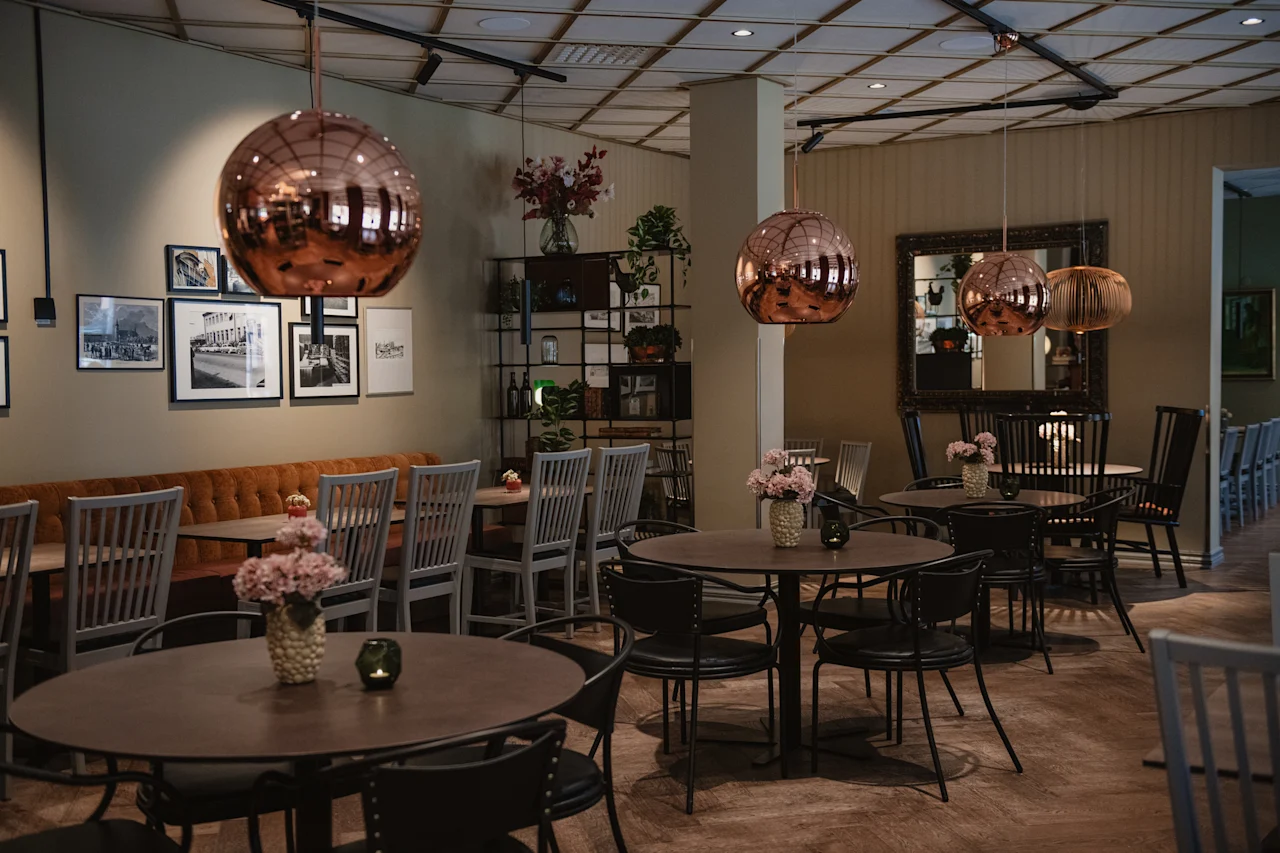 Et varmt og innbydende restaurant- eller kaféinteriør med bord, stoler, en lang benk, kobberpendellamper og innrammet kunst.