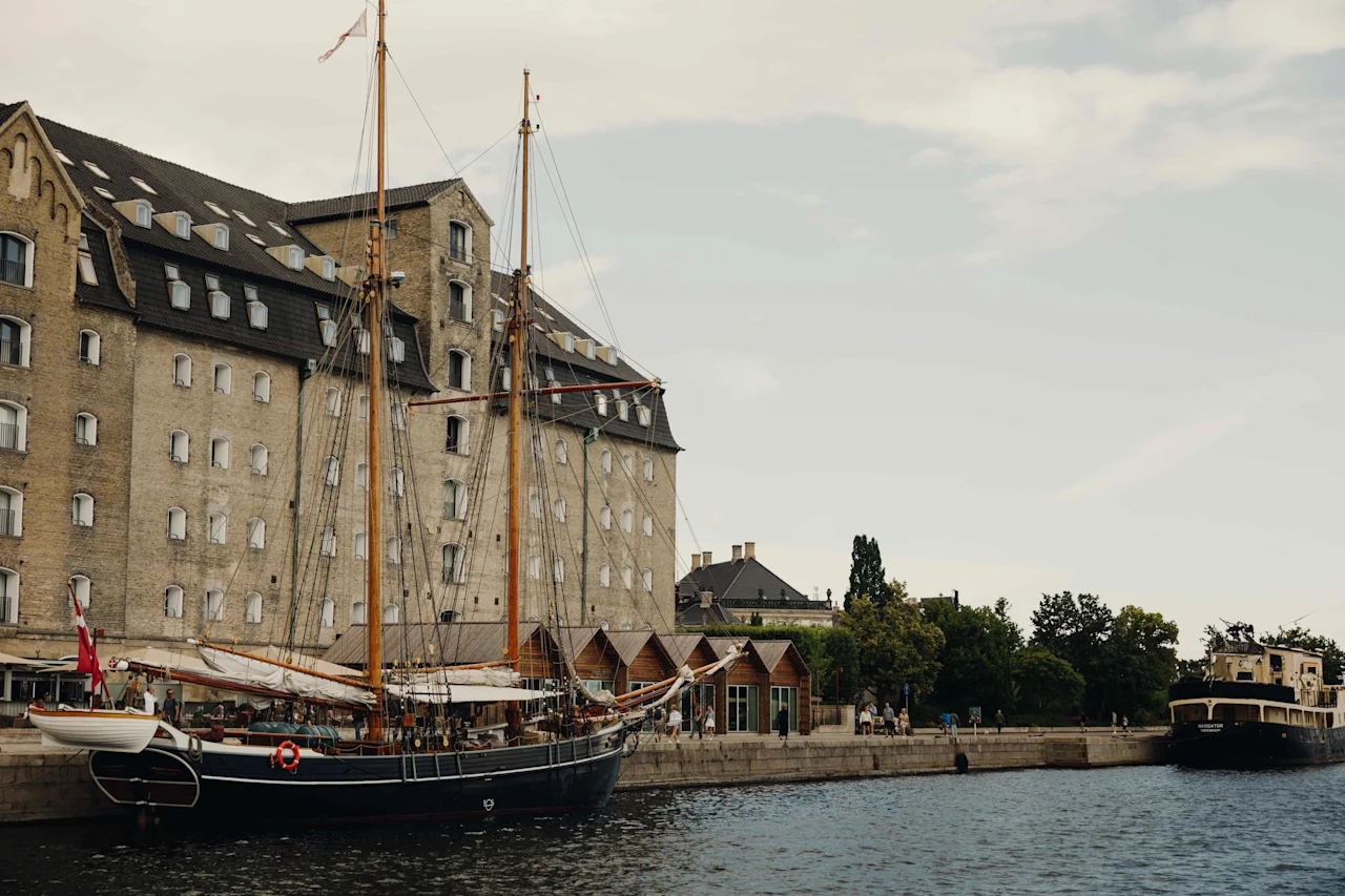 En historisk kanalscene med stor mursteinsbygning, seilbåt fortøyd og annen båt i det fjerne. Folk langs vannkanten.