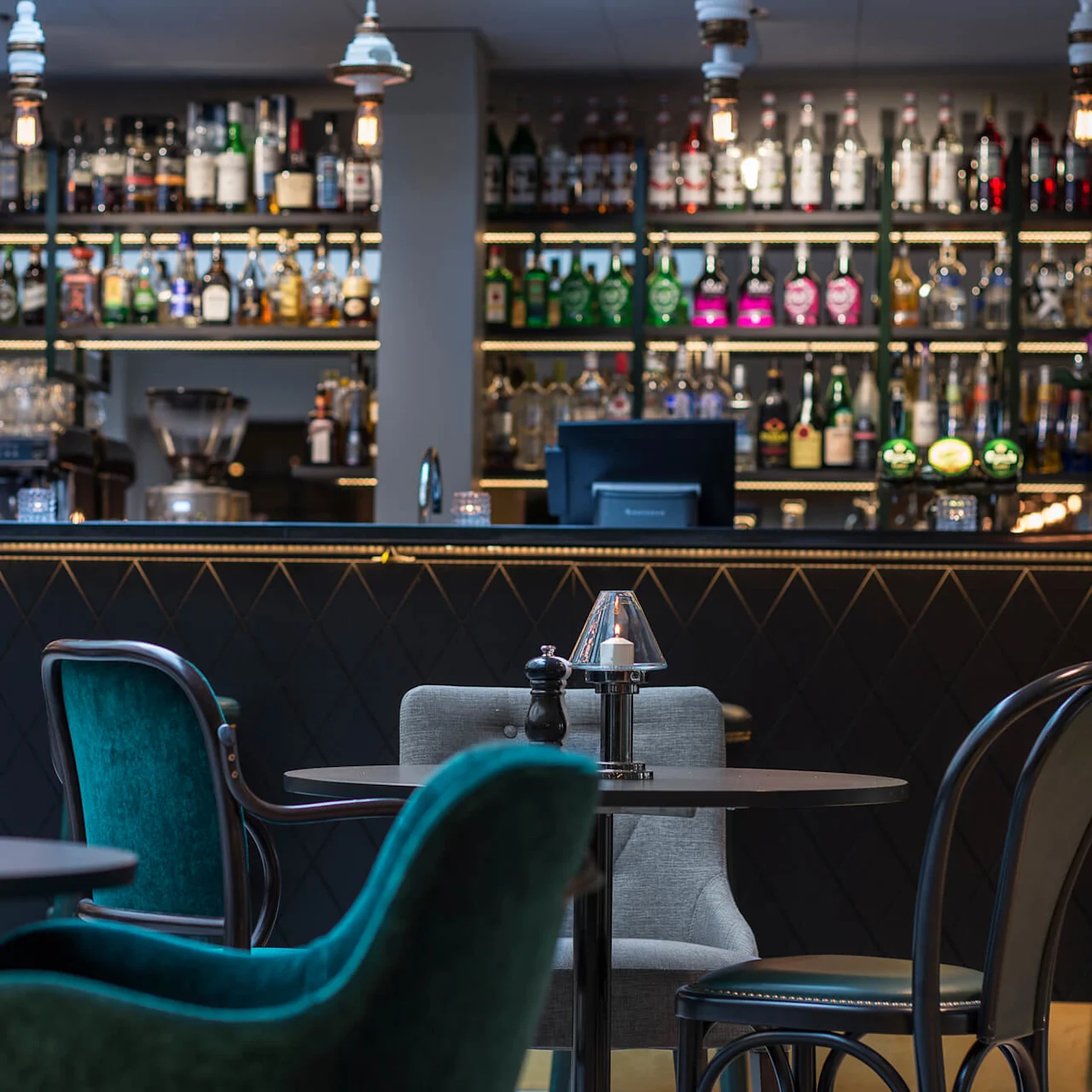 Bord med stolar arrangerade i en svagt upplyst bar, med en välfylld sprithylla i bakgrunden som framhäver den eleganta och sofistikerade miljön.