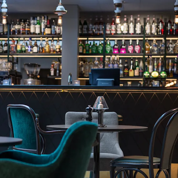 Bord med stolar arrangerade i en svagt upplyst bar, med en välfylld sprithylla i bakgrunden som framhäver den eleganta och sofistikerade miljön.