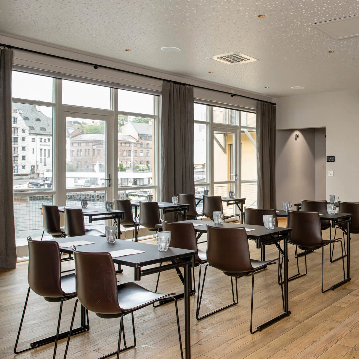 Rows of black chairs and tables face a white projector screen in a conference room with large windows and wooden floors.