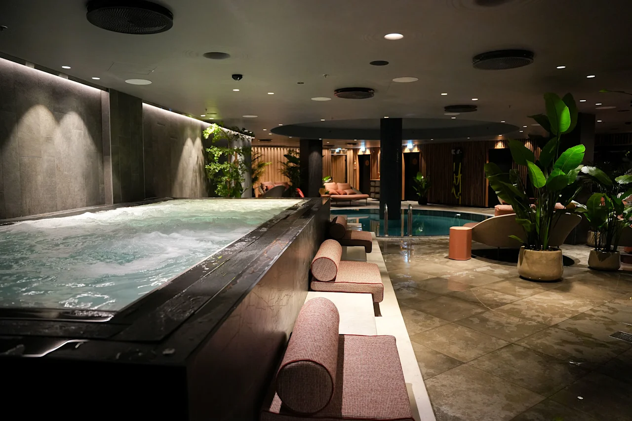 A luxurious indoor spa area with a large bubbling hot tub, lounge chairs, and a serene swimming pool in the background.