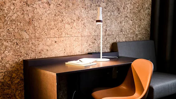 A desk holds a book and lamp, with a brown chair in front. The setting is a cozy room with cork-paneled walls and a gray sofa. 