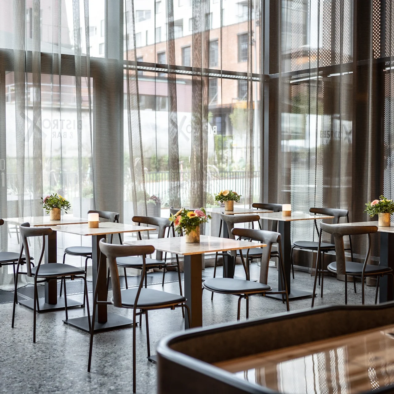 Bord med stolar är arrangerade i ett modernt café, med små blomarrangemang och ljus. Stora fönster i glas ger naturligt ljus, och byggnader syns utanför.