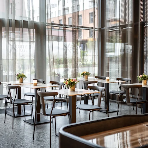 Bord med stoler er arrangert i en moderne kafé, med små blomsterdekorasjoner og lys. Store glassvinduer gir naturlig lys, med bygninger synlige utenfor.