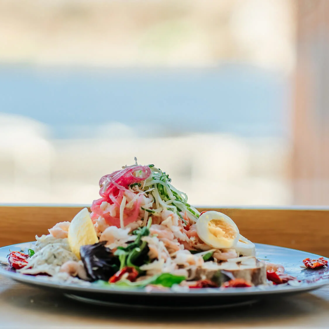 A plate of salad with sliced eggs, shredded meat, and vegetables stacked high, placed on a wooden table with a blurred outdoor background.