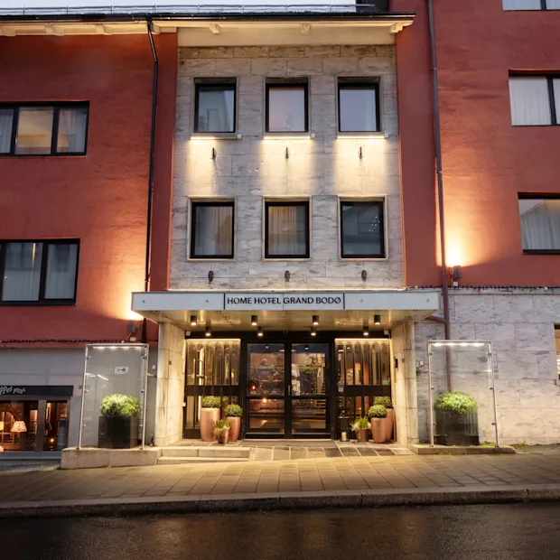 The illuminated entrance of a hotel building at dusk, with the sign HOME HOTEL GRAND BODO above the doors.