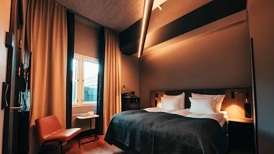 Et hotellrom har en dobbeltseng med hvite puter og mørkt sengetøy. Varm belysning, et vindu med gardiner, en rød stol og et lite bord gir en koselig atmosfære.