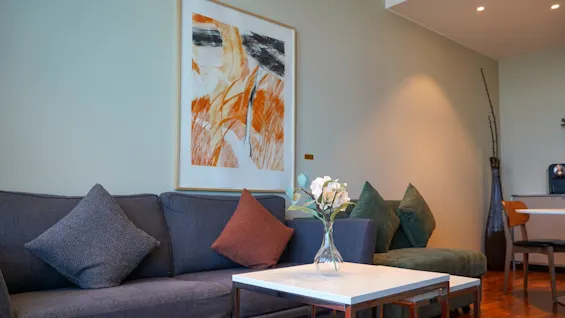 A gray sofa with colorful pillows sits in a modern living room. A vase with white flowers on a white coffee table. Abstract artwork hangs on the wall.