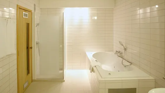 A white-tiled bathroom features a large bathtub and a walk-in shower with frosted glass, adjacent to a wooden sauna door. Bright, ambient lighting fills the room.