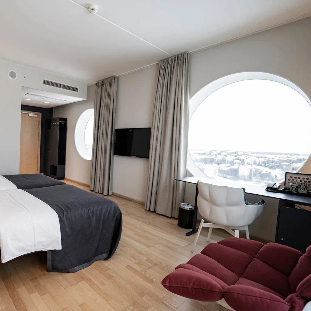 A modern hotel room features two neatly made beds with gray covers, situated next to a large circular window with a cityscape view. A desk and chair are nearby.