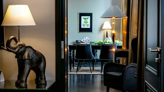 Elefant-formet lampe står på et bord, lyser opp en korridor som leder til en spisestue med et dekket bord. Et innrammet kunstverk og flere lamper pynter rommet. 