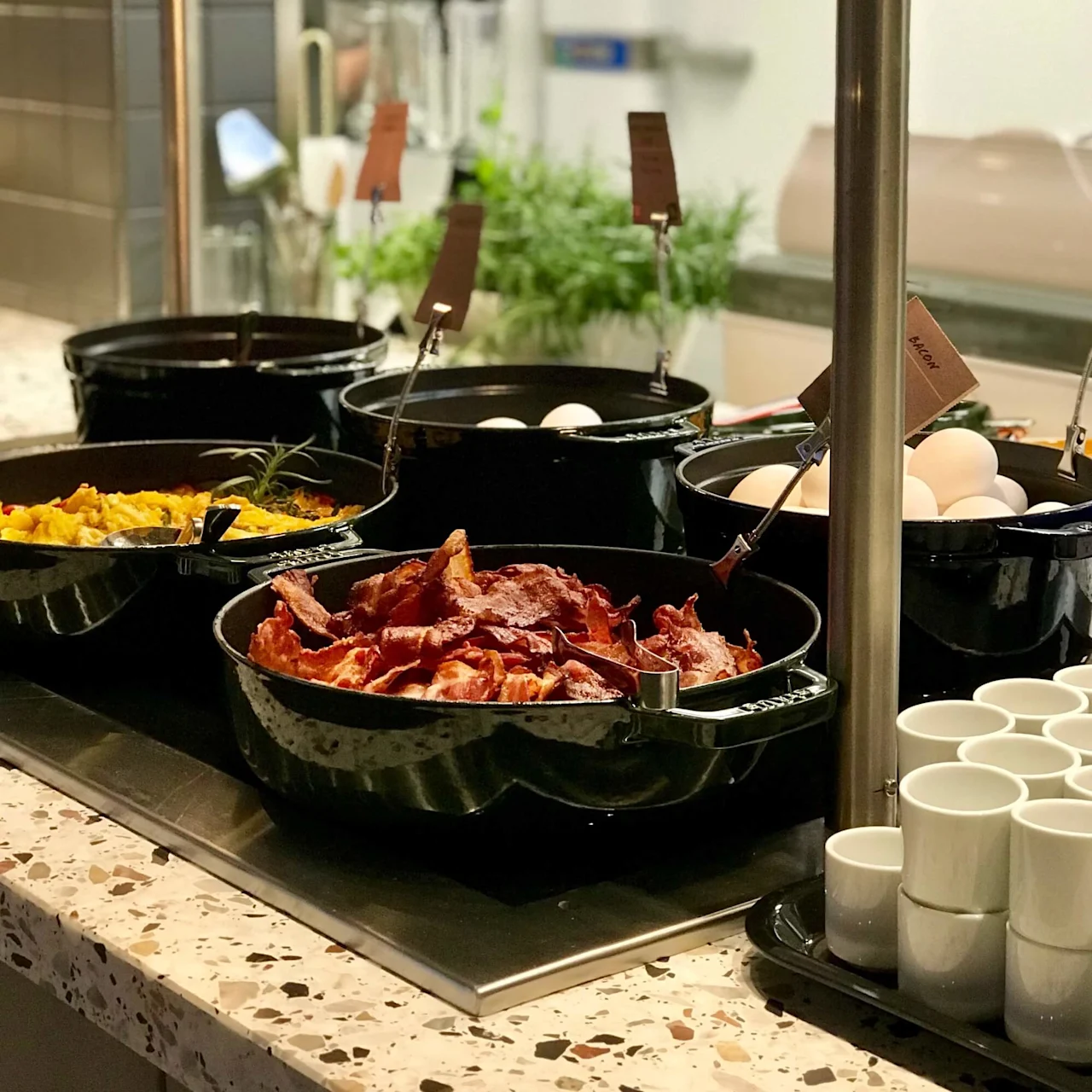 Buffetstation med fade med røræg, bacon og hele æg, placeret på en bordplade omgivet af små hvide skåle og grønne potteplanter.