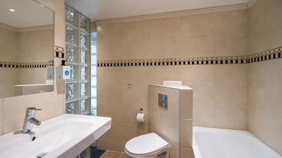 Sink reflecting in mirror, wall-mounted toilet with paper holder, bathtub on tiled floor; beige tiles with decorative band; recessed lighting brightens the modern, compact bathroom.