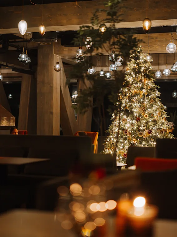 Juletre dekorert med lys og ornamenter, stående i en koselig opplyst restaurant med stearinlys på bordene. Hengende lys skaper en varm og innbydende atmosfære.