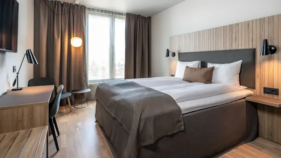 Et pænt indrettet hotelværelse har en stor seng med hvide lagener og brune detaljer, flankeret af moderne lamper. Et skrivebord med stol står i nærheden, med bløde gardiner ved et vindue.