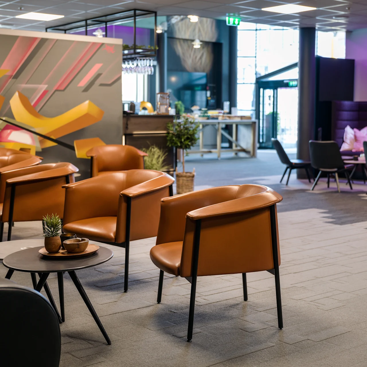 Chairs arranged around tables in a modern lounge with colorful wall art. Additional seating is visible, along with a counter and greenery in the background. No text is present.