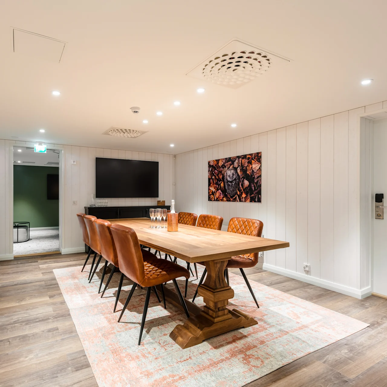 Long wooden table surrounded by leather chairs in a modern conference room, with a TV on the wall and a sign reading "Staff Only" on a door.
