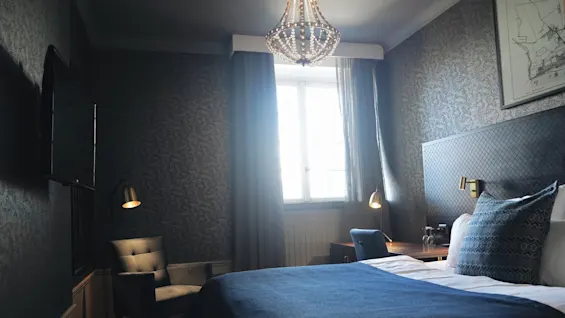 En seng med blått sengetøy står i et stilfullt soverom. Et stort vindu lyser opp rommet, som har en stol, et skrivebord, en lysekrone og dekorativ tapet. En TV er montert på veggen.