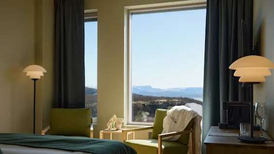 A cozy hotel room features two green chairs by a window, offering a scenic mountain view. A bathrobe with "HFJORD" hangs on a chair, beside a small table and decorative lamp.