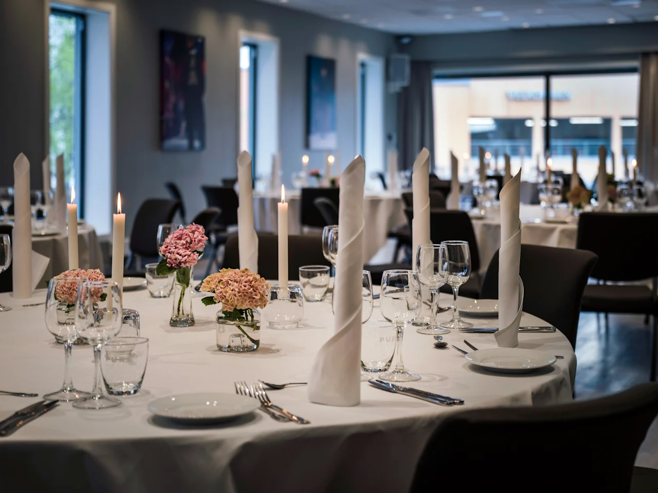 An elegant dining room with round tables set for a formal event, featuring white tablecloths, candles, and floral centerpieces.