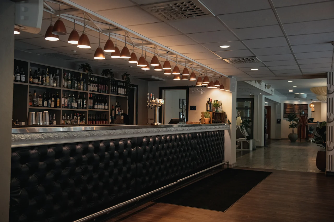 A stylish bar area with a long, dark tufted counter, shelves of bottles, and warm pendant lighting, leading to an open lounge.