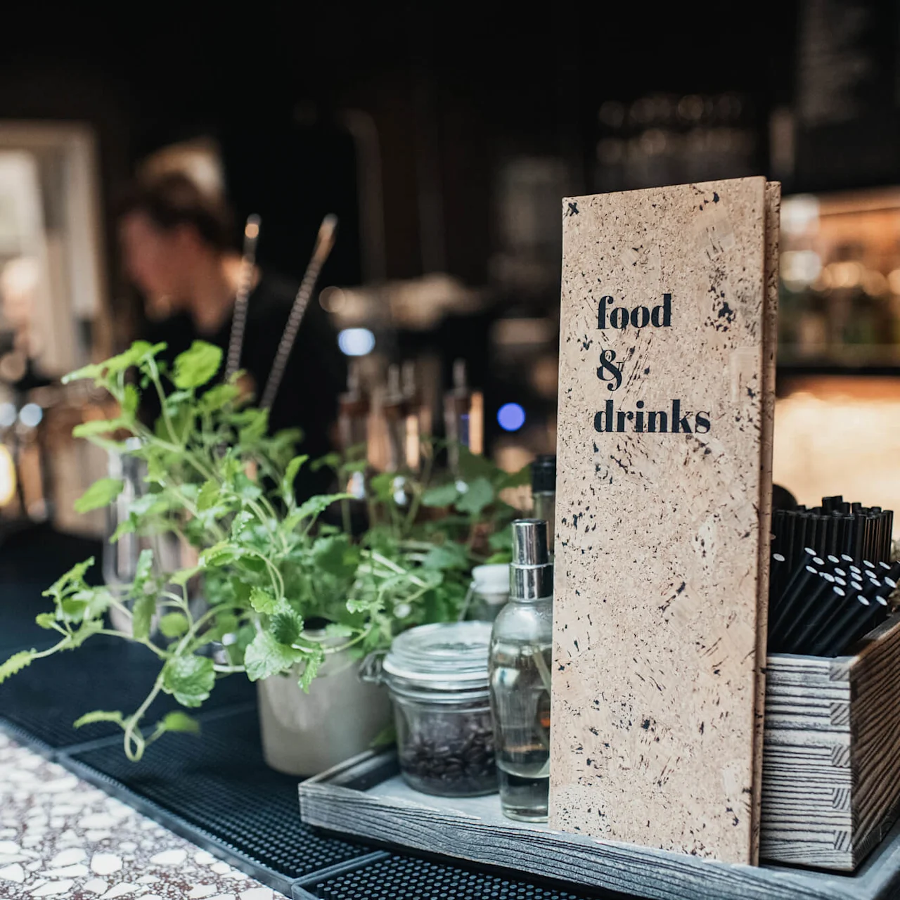A menu with "food & drinks" text stands on a bar counter; blurred bartender in the background.