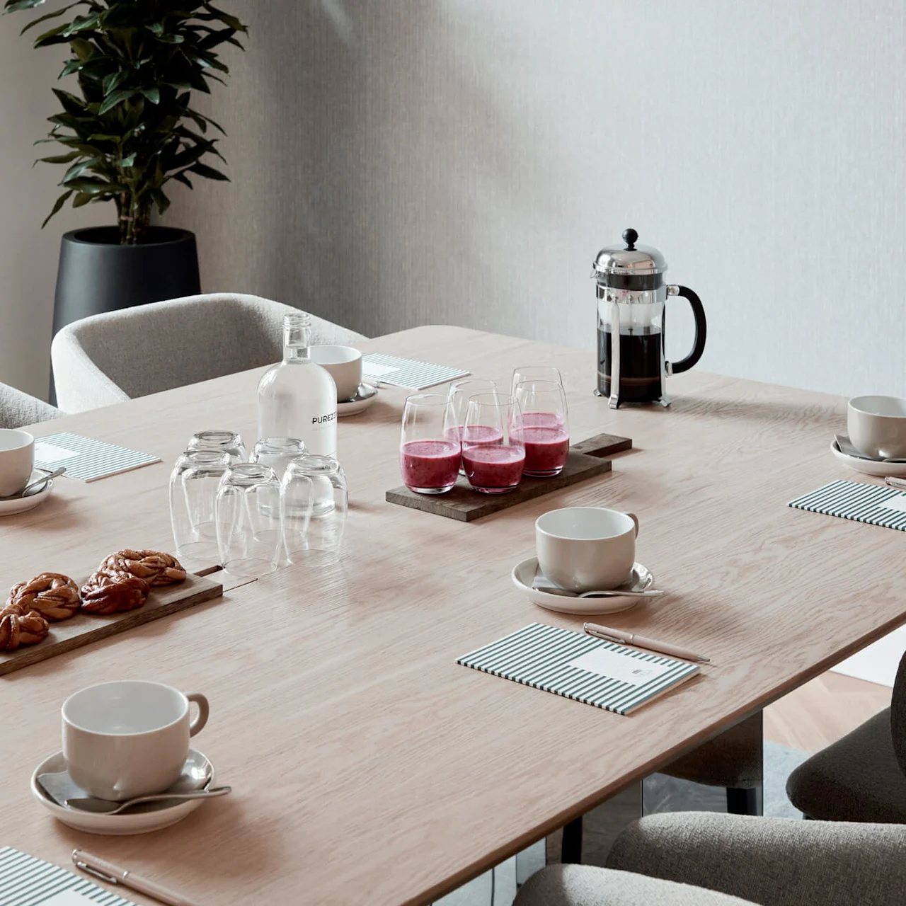 Elegant conference table set for meeting with cups, pastries, and drinks. Potted plant in background. Bottles labeled "PUREZZA".