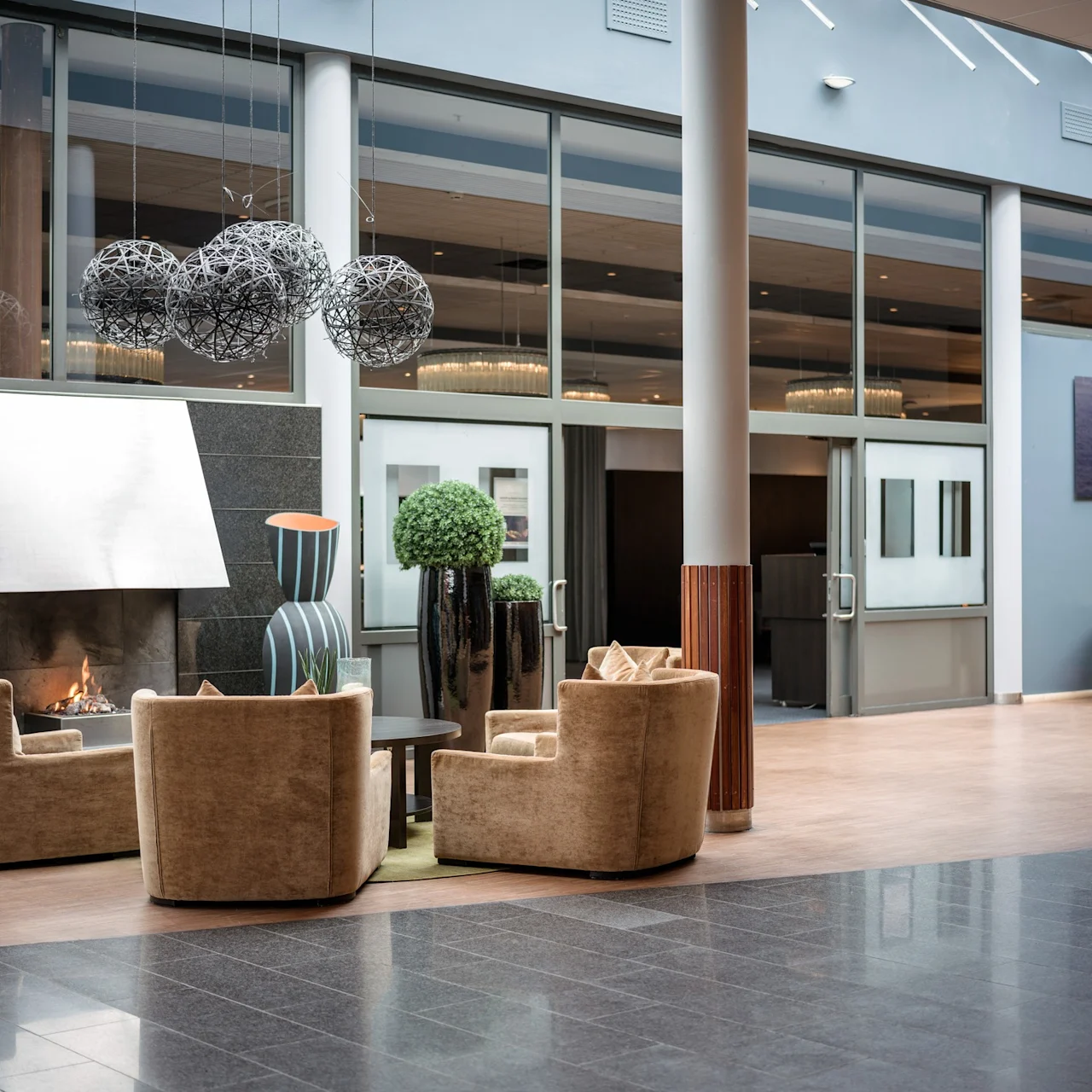 Sittgrupp med fyra beige fåtöljer och ett lågt bord runt en öppen spis; modern konst och vaser pryder hotellobbyn med glaspartier och mjuk belysning.