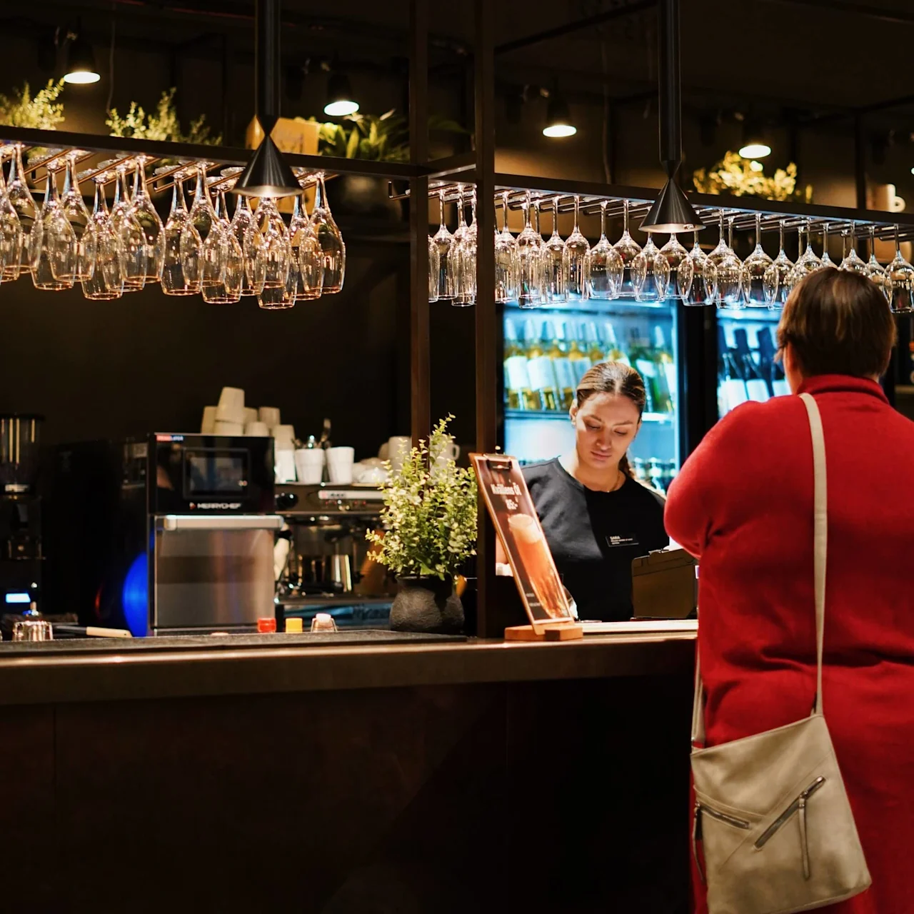 A woman in a red coat stands at a bar counter speaking to a bartender. Wine glasses hang overhead, and beverage coolers are visible in the bar's dimly lit interior.