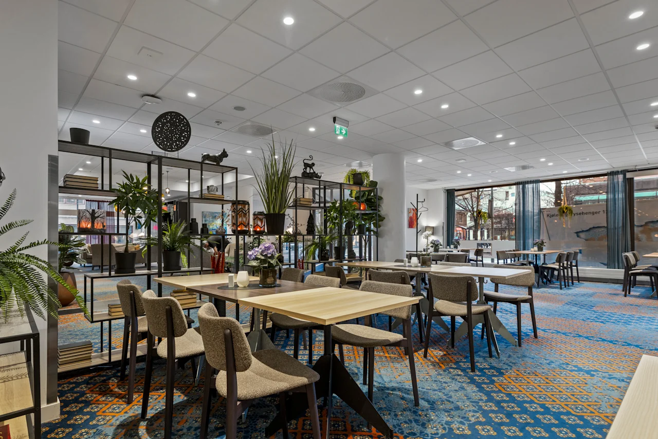 En rymlig matsal på hotell med flera bord och stolar, dekorerad med växter och hyllor. Stora fönster vetter mot en gata. Kjole rsebenger 1.