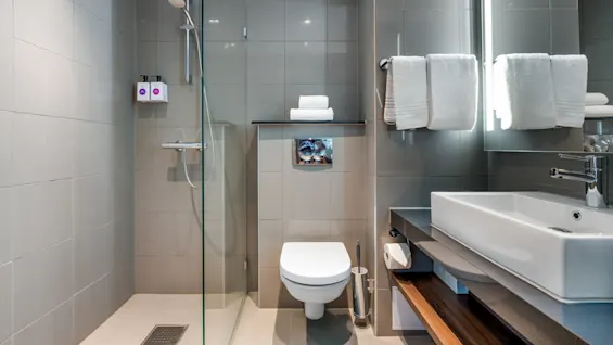 A modern bathroom with tiled walls, glass shower, towels on a rack, and a sink. Neutral color scheme.