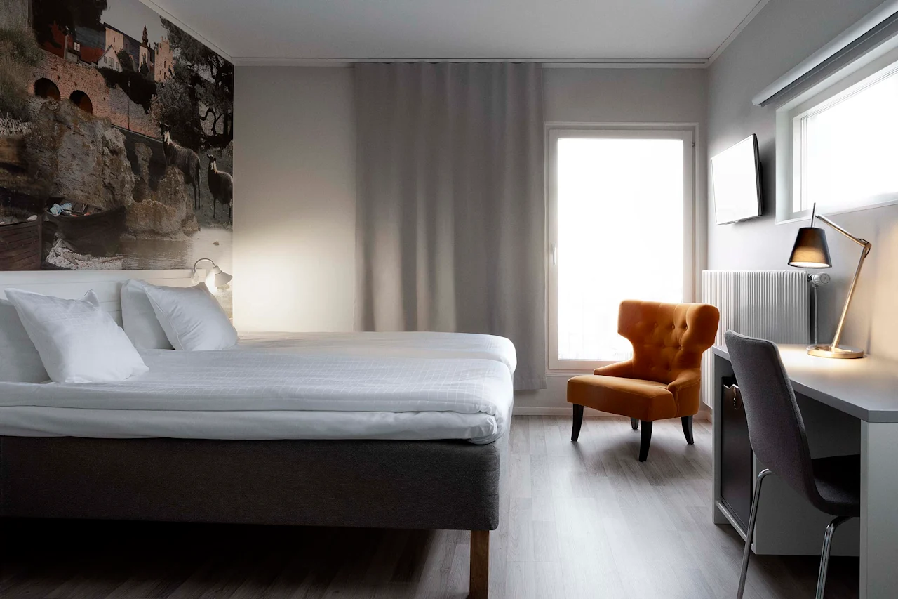 Ett ljust och inbjudande hotellrum med en bekväm säng, en mysig fåtölj och en funktionell skrivbordsplats.