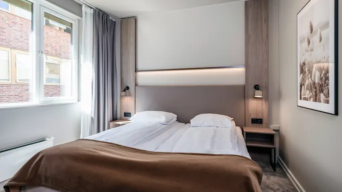 A neatly made double bed with white linens and a brown blanket, flanked by wooden side panels with lamps, in a bright room with a large window and framed wall art.