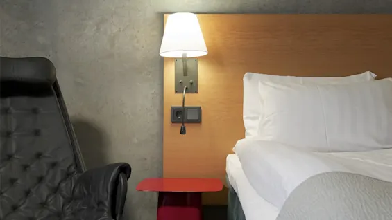Bedside lamp illuminates a wooden headboard. Adjacent, a black leather chair is positioned beside a red table. The room has a concrete wall and dark wooden floor.