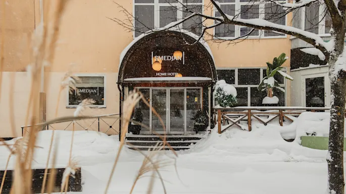 En snefyldt vinterscene uden for en hotelindgang med et 'HOME HOTEL'-skilt. Sne dækker jorden og træerne.