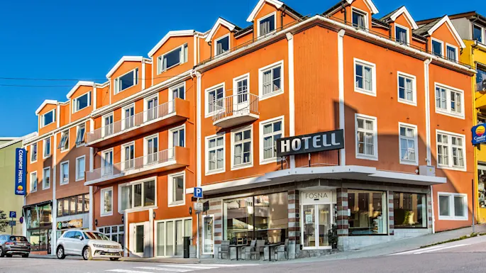 A multi-story orange hotel with large windows and balconies displays signs: "HOTELL," "Comfort Hotel," and "FOSNA." Parked cars are in front, with clear blue sky above.