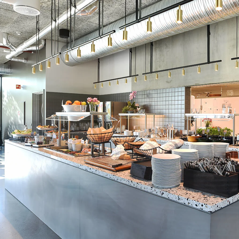 En buffetdisk viser frugt, tallerkener og bestik i en moderne restaurant med industriel indretning og belysning ovenover.