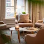 Et koselig stueoppsett har fire lenestoler rundt et rundt trebord med et sjakkbrett og et stearinlys, innrammet av et vindu med gardiner og en gulvlampe.