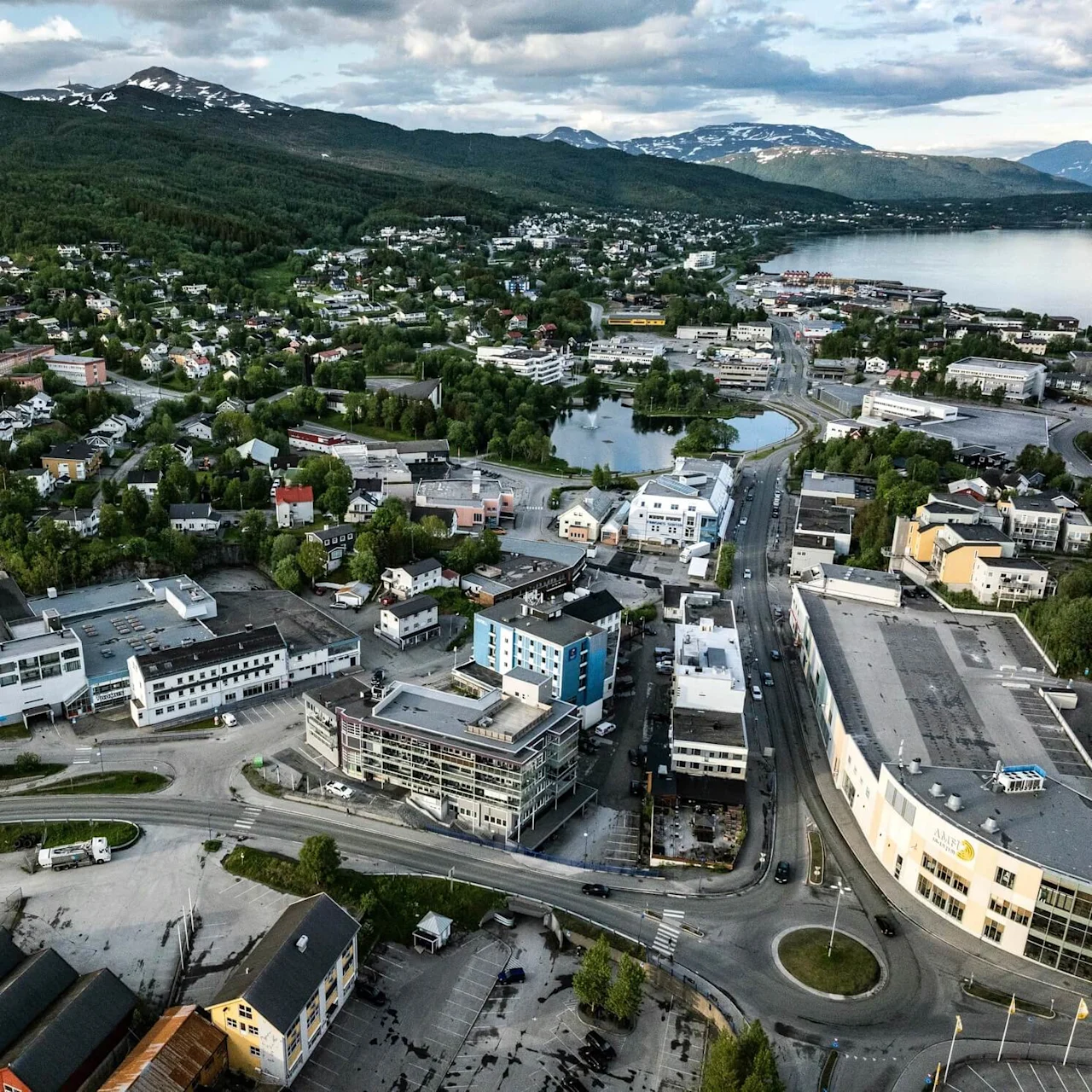 Aerial view of a small town with scattered buildings and greenery, nestled between a lake on the right and mountains in the background beneath a partly cloudy sky.