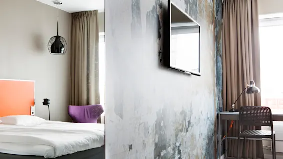 Et vægmonteret TV vender mod et rum med en seng, lilla stol og skrivebord; moderne indretning med neutrale vægge og gardiner skaber en moderne atmosfære.
