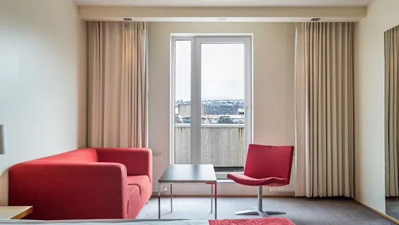 Punainen sohva ja tuoli ovat vastakkain pienen pöydän ääressä hotellihuoneessa. Isot ikkunat verhoilla tarjoavat näkymän kaupunkimaisemaan, kehystettynä vaaleilla seinillä.