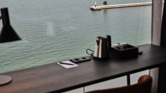 Skrivebord med lampe og elkedel ser ud over en rolig sø og fjerne bakker, set gennem et stort vindue.