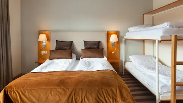 Kaksi vuode, ruskeat tyynyt ja peitot, on seinää vasten kahden seinälampun kanssa. Vieressä on valkoiset kerrossängyt hotellihuoneessa, jossa on matto.